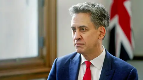 Ed Miliband pictured during his interview. He has short, dark, greying hair. He is wearing a navy suit, white shirt and red tie. Behind him is a wooden window frame and a Union Jack flag.