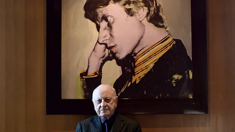 Stephane de Sakutin/AFP/Getty Images Saint Laurent’s partner Pierre Bergé poses in front of an Andy Warhol portrait of the designer (Credit: Stephane de Sakutin/AFP/Getty Images)